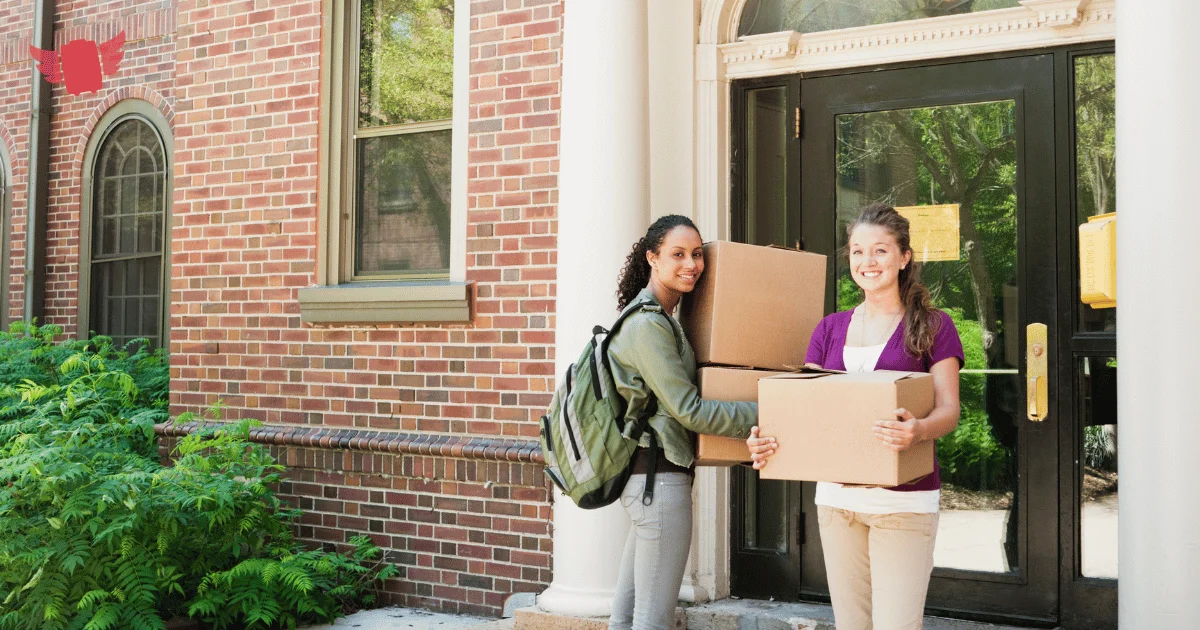 Two students carrying moving boxes up to a red-brick campus dorm on move-in day